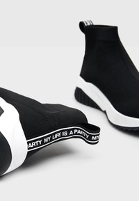 Sneaker nere in stile calza con texture a coste, suola bianca e una linguetta con il testo "LA MIA VITA È UNA FESTA" in bianco.