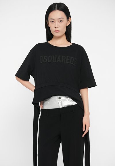 Noor mustaga mustade juuste ja musta DSQUARED2 T-särgiga noor naine, mustade pükste ja hõbedase vööaktsentiga, poseerib valge tausta ees.