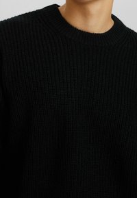 Mężczyzna ubrany w czarny, prążkowany sweter z okrągłym dekoltem, pokazany od szyi do połowy torsu na jednolitym tle.