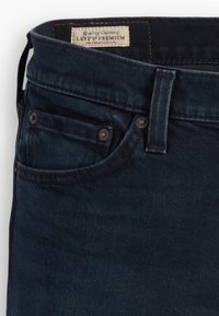 Jean en denim bleu foncé avec poche avant et passants de ceinture, avec une étiquette blanche Levi's Premium cousue à l’intérieur de la ceinture.