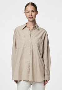 Camicia beige a maniche lunghe con chiusura a bottoni, due tasche sul petto e strisce verticali bianche. Texture liscia e vestibilità morbida.