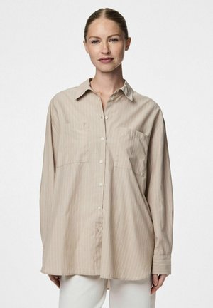 Chemise beige à manches longues avec un devant à boutons, deux poches poitrine et des rayures verticales blanches. Texture lisse et coupe décontractée.