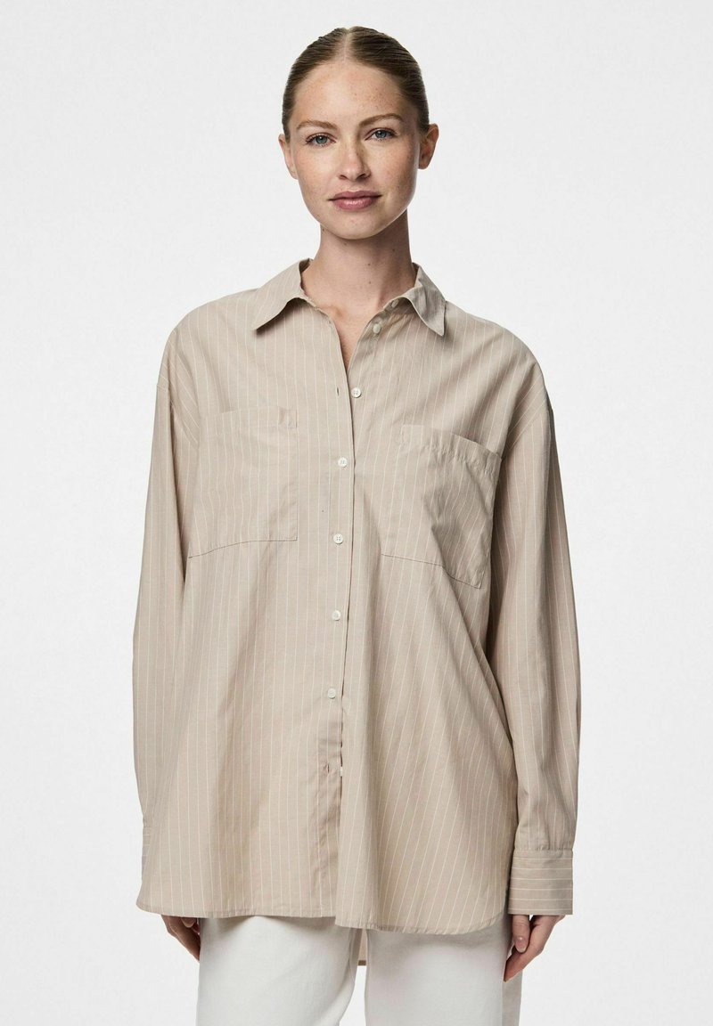 Camicia beige a maniche lunghe con chiusura a bottoni, due tasche sul petto e strisce verticali bianche. Texture liscia e vestibilità morbida.
