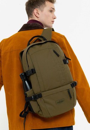 Mochila verde oliva con correas ajustables, múltiples bolsillos y un bolsillo de malla lateral que sostiene una botella de agua negra. Se pueden ver los acentos cosidos.