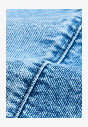 Svijetloplava traper tkanina, s teksturiranim tkanjem i vidljivim šavovima duž rubova, prikazuje različite nijanse plave boje.