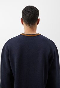 Marinblå stickad tröja med ribbad rund halsringning och en kontrasterande orange-brun detalj. Avslappnad passform med texturerad tyg.