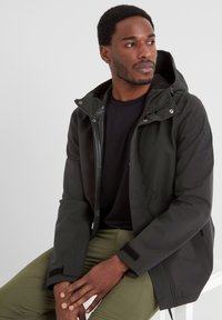 Black waterproof jacket with a zippered front, adjustable hood, and Velcro cuffs. Worn over a black shirt and olive green pants.