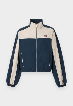 Laivastonsininen ja beige lyhyt takki, jossa on joustavat hihansuut, vetoketjullinen suljenta, sivutaskut ja brändilogot vasemmassa rinnassa. Sileä kangaspinta.