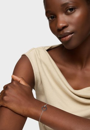 Donna dalla pelle scura che indossa un top beige senza maniche e un delicato braccialetto d'argento con un ciondolo circolare al polso, su uno sfondo neutro.