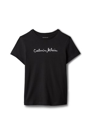 Junges Mädchen mit langen Haaren, das ein schwarzes Calvin Klein T-Shirt, einen blauen Jeansrock und schwarze Sandalen trägt, steht vor einem grauen Hintergrund.