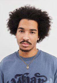 Dunkappet genser med rund hals i mørkeblått med broderte tekstdetaljer. Gullkjede med anheng, kort krøllete svart hår, nøytral bakgrunn.