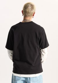 Camiseta de algodón negra con cuello redondo y mangas cortas, llevada sobre una camisa de manga larga blanca. Detalles de costura visible y corte relajado.