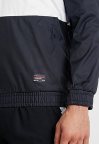 Zwart-witte windbreaker jas met elastische manchetten en zoom, voorzien van een logolabel aan de zijkant. Soepele textuur en lichtgewicht materiaal.
