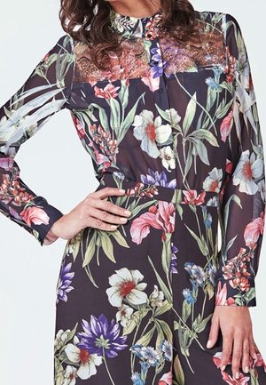 Kobieta ubrana w przezroczystą, długorękawową sukienkę w kwiaty z zapięciem na guziki z przodu oraz koronkowymi detalami na górnej części klatki piersiowej i ramionach.