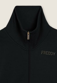 Chaqueta negra con cremallera y cuello alto, con el logo dorado con tachuelas "FREDDY" en el lado izquierdo del pecho, que cuenta con un tirador de cremallera metálico marcado con "FREDDY".