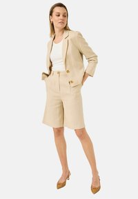 Beige linned blazer med to guldknapper, parret med matchende shorts til midt på låret. En hvid top og brune vævede stilethæle fuldender looket.
