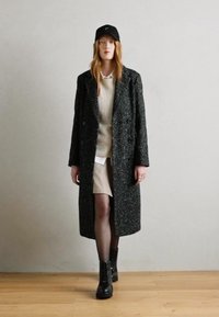 Long manteau sombre texturé avec des boutons double boutonnage, porté sur un pull léger et une jupe beige, complété par des bottes à lacets noires et une casquette.