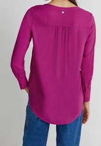 Blusa magenta a maniche lunghe con orlo arrotondato, caratterizzata da una piega sul retro e un piccolo bottone sul collo. Tessuto morbido e leggero.