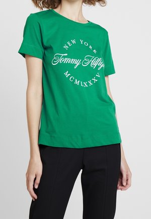 Vrouw draagt een groene Tommy Hilfiger T-shirt met "New York" en witte Romeinse cijfers, gecombineerd met een zwarte broek.