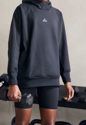 Osoba ubrana w czarną bluzę Adidas i szorty, trzymająca hantle na siłowni, z ciężarami na stojaku w tle.