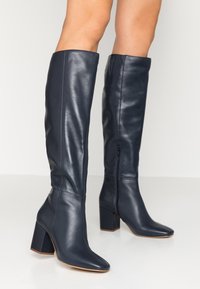 Bottes en cuir bleu marine montant jusqu'au genou, avec un talon bloc, un bout carré, et une fermeture éclair latérale. Texture lisse et design ajusté pour une apparence épurée.