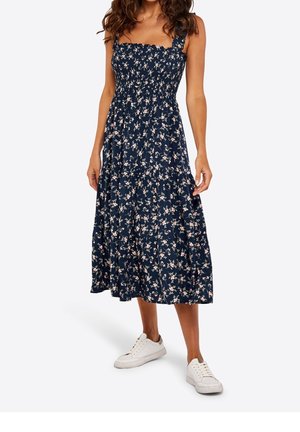Femme portant une robe midi bleu marine avec un petit motif floral et des baskets blanches, debout devant un fond uni.