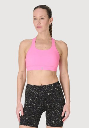 Vrouw met gevlochten haar, die een roze sportbeha en zwarte, hoog uitgesneden fietsbroek met spikkels draagt, staand tegen een lichtgrijze achtergrond.