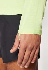 Langärmliges, hellgrünes Sportoberteil mit Daumenlöchern, kombiniert mit dunklen Shorts, das eine glatte Textur und eine enge Passform aufweist.