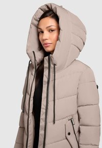 Beige pufferjack met een grote capuchon, frontverener en zijzakken. Heeft horizontale stiksels en zwarte koordjes.
