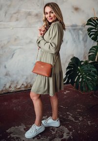 Oliven grünes Kleid mit einem ausgestellten Rock, braune Umhängetasche mit goldener Kette und weiße Sneakers mit metallischen Akzenten. Grüne Pflanze im Hintergrund.