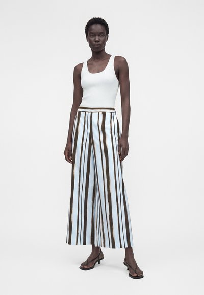 Γυναίκα που φοράει λευκό τοπ χωρίς μανίκια, παντελόνι με φαρδιά μπατζάκια με κάθετες καφέ και μπλε ρίγες και μαύρα σανδάλια με τακούνι, στέκεται μπροστά σε απλό φόντο.