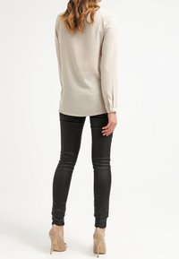 Blouse beige à manches longues avec une texture lisse, associée à un jean skinny noir et des talons hauts couleur nude. La blouse est dotée de manches ajustées.