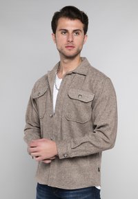 Bruine overhemdjack met visgraatpatroon, twee borstzakken, knoopsluiting en een kraag, gecombineerd met een witte T-shirt.