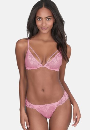 Woman with long dark hair wearing pink lace bra and matching lace panties, standing against a plain light background.