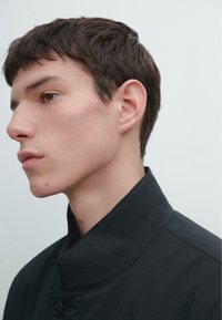 Sort jakke med høj krave, glat tekstur og knaplukning. Har rene linjer og minimalistisk design. Nærbillede profiludsigt.