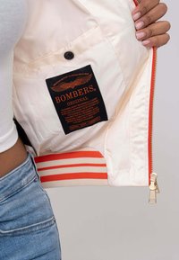 Crème bomberjack interieur met zwart en oranje label; voorzien van rood gestreepte elastiek aan de onderkant en een gouden rits.