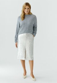 Vrouw staand in een grijze trui, metallic zilveren knielange rok met split en crèmekleurige pumps met spitse neus tegen een eenvoudige lichte achtergrond.