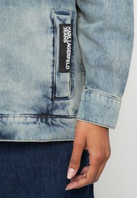 Svetlo modra denim jakna s zbledelo teksturo, na kateri je črna znamkasta oznaka. Manšeta prikazana z vidnim šivom.