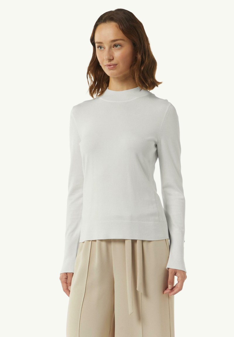 Witte, lange mouwen coltrui van gladde stof, met ribbels aan de mouwen en onderkant, gecombineerd met lichtbeige broeken.