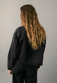 Čierna rifľová bunda s voľným strihom a klesnutými ramenami, kombinovaná s čiernymi džínsami rovnakej farby. Vlasmi stočenými do kučier padajúcimi po chrbte.