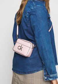 Kobieta w niebieskiej dżinsowej kurtce i kratkowanej spódnicy, niosąca małą różową torebkę Calvin Klein na ramię z srebrnym łańcuszkiem i logo.