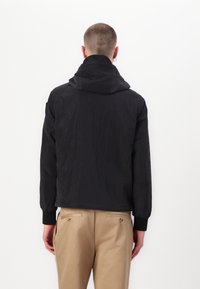 Black hooded jacket with a textured fabric and elastic cuffs. Displayed from the back, it features a minimalist design and a cropped fit.