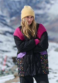 Čierna prešívaná bunda s fialovými a kvetinovými detailmi, ružové textúrované rukávy a žltá pletená čiapka. V pozadí je vidieť zasneženú krajinu.