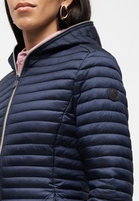 Γυναίκα που φοράει σκούρο μπλε μπουφάν καπιτονέ με κουκούλα, χρυσό φερμουάρ και στρογγυλό λογότυπο-μπατζάκι στο επάνω μέρος του βραχίονα, συνδυασμένο με χρυσούς κρίκους σκουλαρίκια.