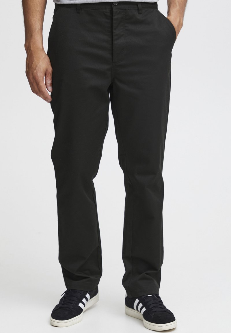 Black cotton-blend trousers with a straight-leg design, flat front, and side pockets, paired with black sneakers featuring white stripes.