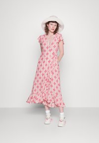 Vestido envolvente floral rosa com mangas e bainha onduladas, apresentando um detalhe de laço na cintura; modelo combinado com ténis brancos e um chapéu de aba larga.