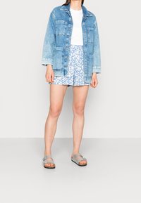Jasnoniebieska kurtka dżinsowa z czterema kieszeniami i kołnierzem, zestawiona z jasnoniebieskimi szortami w kwiaty. Szare sandały z regulowanymi paskami.