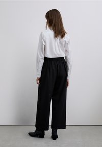 Camisa blanca de manga larga con puños abotonados combinada con pantalones negros de pierna ancha con un sutil patrón de rayas finas, usados con botas negras hasta el tobillo.