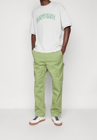 Jasnoszara bluza z napisem "CONVERSE" w zielonym kolorze, zestawiona z jasnooliwkowymi spodniami cargo i białymi trampkami z niebieskimi akcentami.