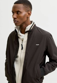 Νέος άνδρας με κοντά μαλλιά, φορώντας σκούρο σακάκι πάνω από ανοιχτό πουλόβερ με φερμουάρ, στέκεται με τα χέρια στις τσέπες, κοιτάζοντας προς τα αριστερά.